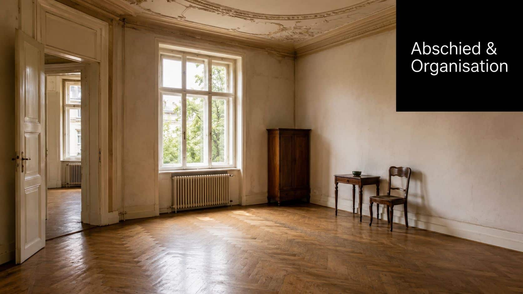 Ein geräumiges, leeres Zimmer mit Fischgrätparkett, einem Fenster, einem alten Holzschrank sowie Tisch und Stuhl.